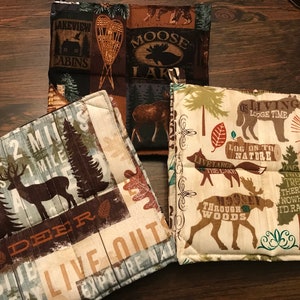 May include: Set of three brown, beige, and green fabric pot holders with a woodland theme. The pot holders feature various designs including deer, moose, trees, and text such as "Lake 2 Miles", "Live Out", "Log On To Nature", and "Through The Woods".
