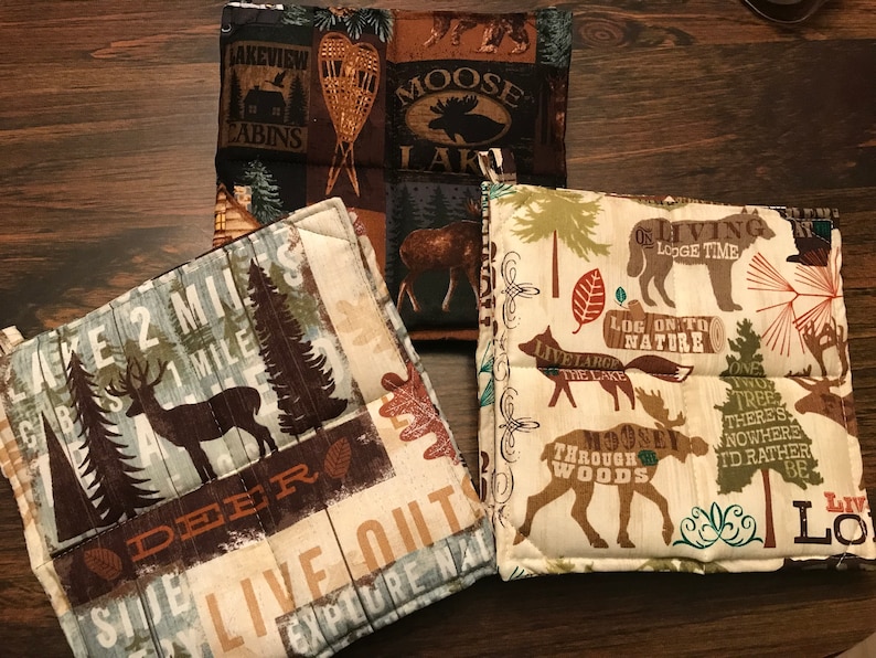 Puede incluir: Conjunto de tres agarraderas de olla de tela marr&oacute;n, beige y verde con un tema de bosque. Las agarraderas presentan varios dise&ntilde;os que incluyen ciervos, alces, &aacute;rboles y texto como "Lake 2 Miles", "Live Out", "Log On To Nature" y "Through The Woods".