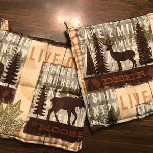 Puede incluir: Dos agarraderas de olla acolchadas de color marr&oacute;n y beige con un tema de bosque. Una agarradera de olla presenta un alce, &aacute;rboles y el texto "Live Out Explore Raft Hear Lake Moose". La otra agarradera de olla presenta un ciervo, &aacute;rboles y el texto "Lake 2 Miles CBS 1 Mile Deer County Live Out Explore Nature".