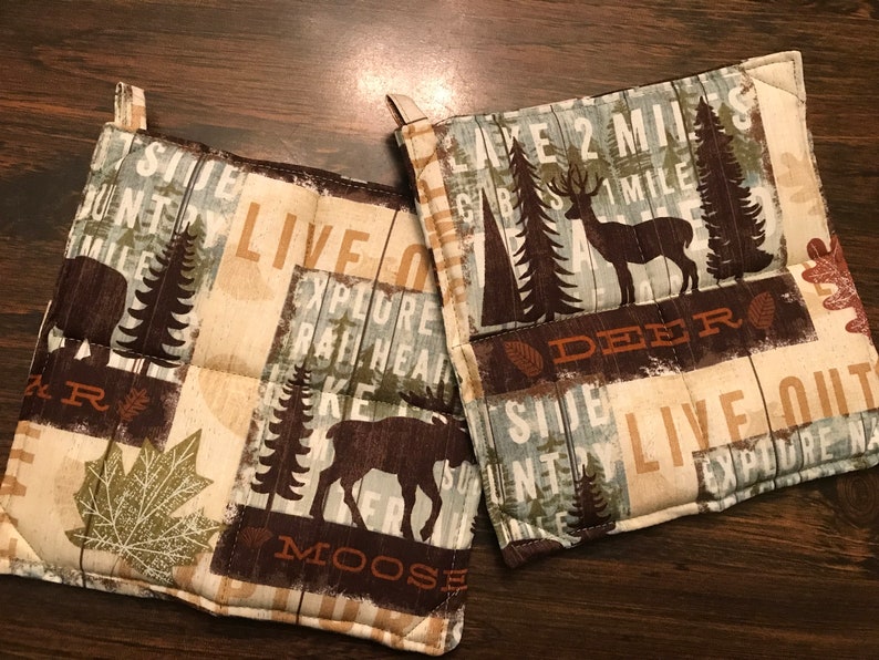 Puede incluir: Dos agarraderas de olla acolchadas de color marr&oacute;n y beige con un tema de bosque. Una agarradera de olla presenta un alce, &aacute;rboles y el texto "Live Out Explore Raft Hear Lake Moose". La otra agarradera de olla presenta un ciervo, &aacute;rboles y el texto "Lake 2 Miles CBS 1 Mile Deer County Live Out Explore Nature".