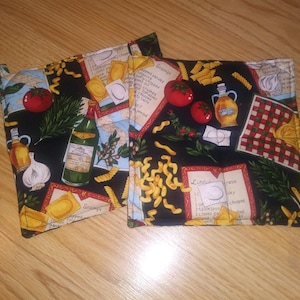 May include: Two black fabric pot holders with a red, yellow, and green Italian-themed design. The design features pasta, tomatoes, garlic, olive oil, and rosemary. The pot holders have a loop handle on each side.