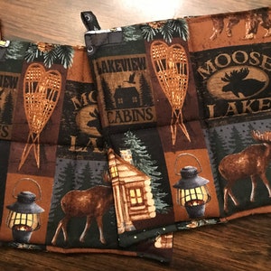 Puede incluir: Conjunto de tres agarraderas de olla acolchadas de color marr&oacute;n y negro con un tema de bosque. Las agarraderas de olla presentan im&aacute;genes de alces, caba&ntilde;as, raquetas de nieve y linternas. Los textos "Lakeview Cabins" y "Moose Lake" est&aacute;n impresos en las agarraderas de olla.