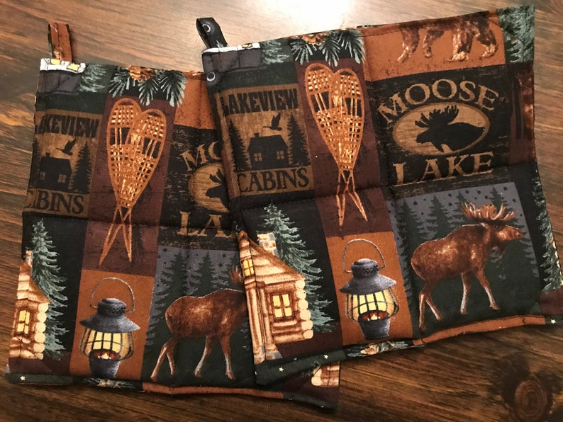 Puede incluir: Conjunto de tres agarraderas de olla acolchadas de color marr&oacute;n y negro con un tema de bosque. Las agarraderas de olla presentan im&aacute;genes de alces, caba&ntilde;as, raquetas de nieve y linternas. Los textos "Lakeview Cabins" y "Moose Lake" est&aacute;n impresos en las agarraderas de olla.
