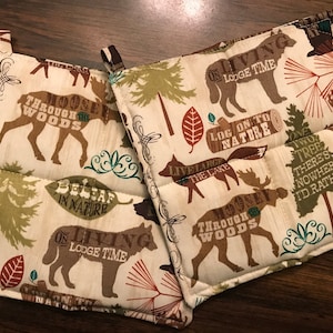 Puede incluir: Dos agarraderas de olla marrones y beige con un tema de bosque. Las agarraderas presentan alces, lobos y &aacute;rboles con texto que dice "Moosey Through the Woods", "On Living Lodge Time", "Log On To Nature", "Live Large The Lake", "Believe In Nature", y "One Two Tree There's Nowhere I'd Rather Be".