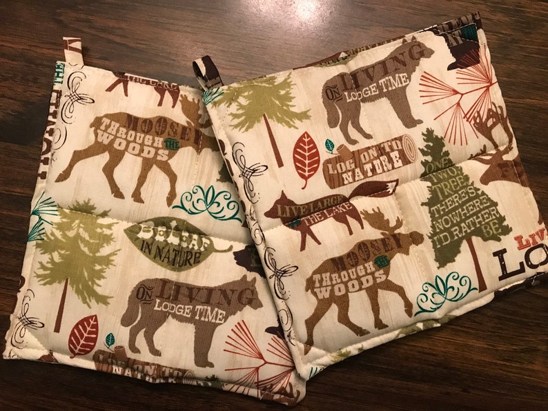 Puede incluir: Dos agarraderas de olla marrones y beige con un tema de bosque. Las agarraderas presentan alces, lobos y &aacute;rboles con texto que dice "Moosey Through the Woods", "On Living Lodge Time", "Log On To Nature", "Live Large The Lake", "Believe In Nature", y "One Two Tree There's Nowhere I'd Rather Be".