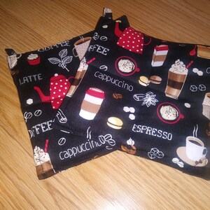 May include: Two black fabric pot holders with a coffee theme. The pot holders have a white background with black and red coffee-themed illustrations and text, including "coffee", "latte", "cappuccino", "espresso", and "coffee beans".