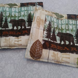 May include: Two quilted pot holders with a nature-themed design. The fabric features a bear silhouette, trees, and the text "LIVE OUTSIDE WILD COUNTRY TALL 1 MILE BEAR" in brown, green, and beige tones. The pot holders are square and have a quilted pattern.