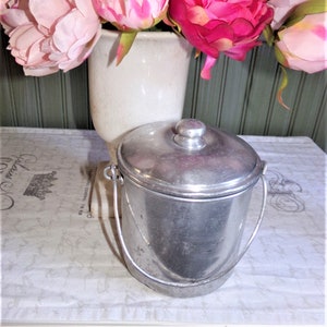 May include: A silver metal container with a lid and a handle. The container is sitting on a white tablecloth with a floral pattern. A bouquet of pink and red peonies is in a white vase behind the container.