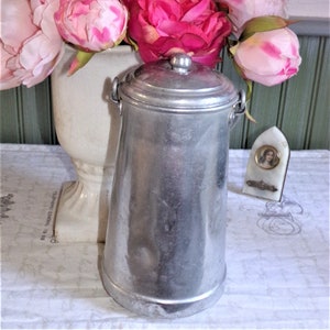 May include: A silver aluminum pitcher with a lid and a handle. The pitcher is sitting on a white tablecloth with a bouquet of pink and white peonies in the background.