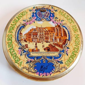 Peut inclure: Boîte métallique ronde vintage avec une illustration détaillée d'une place de ville européenne, avec des bâtiments, une statue et des voitures anciennes. La boîte a un bord doré et un fond jaune avec les mots "VERITABLES PRASLINES" et "MAZET DE MONTARGIS".