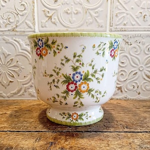 Pode incluir: Um vaso de cerâmica branca com um padrão floral pintado à mão nas cores vermelho, laranja, azul e verde. O recipiente em forma de taça possui uma base de pedestal e uma borda verde, decorado com vinhas e pequenos motivos florais.