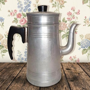 May include: A silver-colored metal coffee percolator featuring a cylindrical body with horizontal embossed lines, a curved spout, and a black heat-resistant handle. The lid is topped with a small, round black knob for easy handling.
