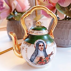 May include: A small porcelain teapot featuring a white base with a green upper section and gold trim. The front displays a detailed illustration of a nun holding a crucifix and roses. It has a gold-colored handle, spout, and lid finial.