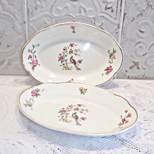 Op de afbeelding: Twee ovale serveerschalen met een witte achtergrond en gouden rand. Elke schaal heeft een bloemmotief met een vogel die op een tak zit.