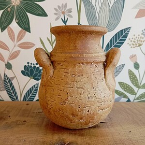 May include: A small, textured, tan-colored ceramic vase with two curved handles. The vase has a rounded body and a flared rim. The background features a floral wallpaper with green, blue, and pink flowers.