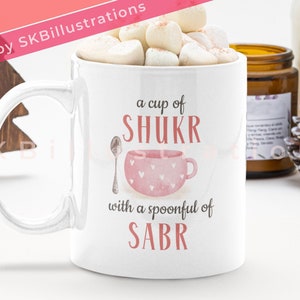 May include: White ceramic mug with a pink and white illustration of a cup and spoon. The text on the mug reads "a cup of SHUKR with a spoonful of SABR".