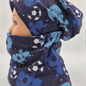 Könnte beinhalten: Ein dunkelblaues Beanie und ein passender Schlauchschal mit einem sich wiederholenden Muster aus weißen Fußbällen und hellblauen Farbspritzern. Der Stoff hat eine glatte Textur und das Design bedeckt beide Teile in einem einheitlichen Druck.