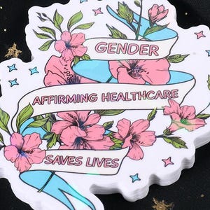 May include: A white sticker with a floral design and the text "GENDER AFFIRMING HEALTHCARE SAVES LIVES". The sticker features pink flowers, green leaves, and a blue ribbon. The background is black with gold stars.