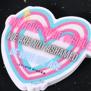 May include: A heart-shaped sticker with a white border and a layered design of pink, blue, and white. The sticker features the text "WE HAVE TO BE VISIBLE WE ARE NOT ASHAMED OF WHO WE ARE -SYLVIA RIVERA."
