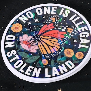 May include: A round sticker with the text "NO ONE IS ILLEGAL" and "NO STOLEN LAND" encircling a butterfly and flowers. The butterfly has orange, black, and white wings. The sticker has a holographic effect.