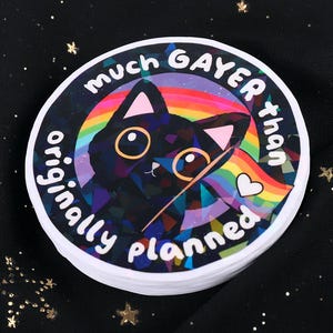 May include: A stack of round stickers with a black cat illustration and rainbow design. The text on the sticker reads "much GAYER than originally planned". The cat has large yellow eyes and a rainbow heart.