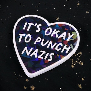 May include: A heart-shaped sticker with the text "IT'S OKAY TO PUNCH NAZIS" in white. The sticker has a white border and a holographic, multi-colored center, set against a black background with gold glitter.
