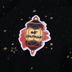 The Holographic lumina converter sticker with the text "We Continue" on top. Behind the keychain is a red paint swipe.