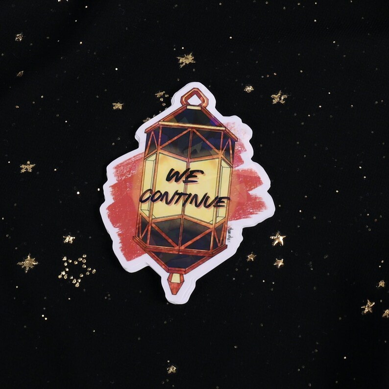 The Holographic lumina converter sticker with the text "We Continue" on top. Behind the keychain is a red paint swipe.