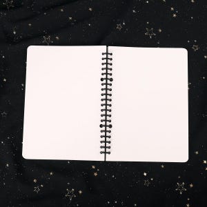 Puede incluir: Un cuaderno blanco de espiral con un patr&oacute;n de cuadr&iacute;cula punteada en las p&aacute;ginas. El cuaderno est&aacute; abierto y acostado sobre un fondo de tela negra con estrellas doradas brillantes.