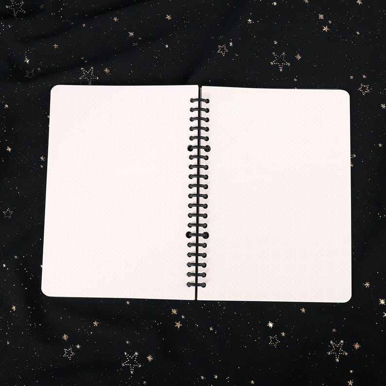 Puede incluir: Un cuaderno blanco de espiral con un patr&oacute;n de cuadr&iacute;cula punteada en las p&aacute;ginas. El cuaderno est&aacute; abierto y acostado sobre un fondo de tela negra con estrellas doradas brillantes.