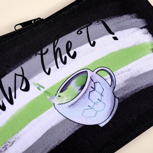 Puede incluir: Estuche negro con cierre de cremallera. El dise&ntilde;o presenta una taza de t&eacute; estilizada con l&iacute;quido verde saliendo y el texto "Us the T?" en una fuente manuscrita. El fondo tiene un dise&ntilde;o de pinceladas en negro, blanco y verde.