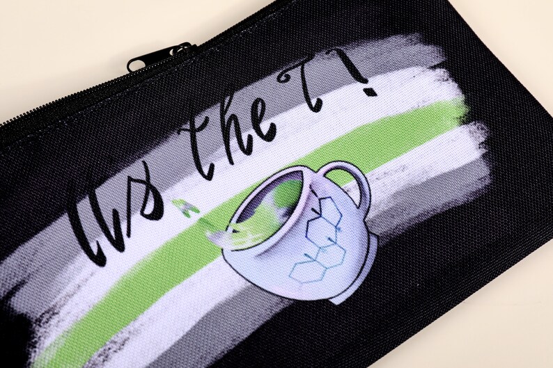 Puede incluir: Estuche negro con cierre de cremallera. El dise&ntilde;o presenta una taza de t&eacute; estilizada con l&iacute;quido verde saliendo y el texto "Us the T?" en una fuente manuscrita. El fondo tiene un dise&ntilde;o de pinceladas en negro, blanco y verde.