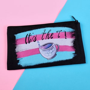 Puede incluir: Bolsa negra con cremallera con el texto "It's the T!" sobre una ilustraci&oacute;n de taza de t&eacute;. El dise&ntilde;o presenta los colores de la bandera del orgullo transg&eacute;nero: azul claro, rosa y blanco. La taza de t&eacute; tiene cubitos de hielo y un l&iacute;quido saliendo.