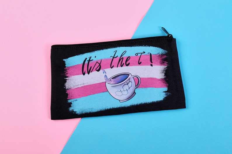 Puede incluir: Bolsa negra con cremallera con el texto "It's the T!" sobre una ilustraci&oacute;n de taza de t&eacute;. El dise&ntilde;o presenta los colores de la bandera del orgullo transg&eacute;nero: azul claro, rosa y blanco. La taza de t&eacute; tiene cubitos de hielo y un l&iacute;quido saliendo.