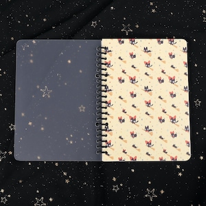 Puede incluir: Un cuaderno de espiral con una cubierta de pl&aacute;stico transparente y una cubierta amarilla con un patr&oacute;n repetido de gatos negros montando escobas. El cuaderno tiene una encuadernaci&oacute;n en espiral negra.