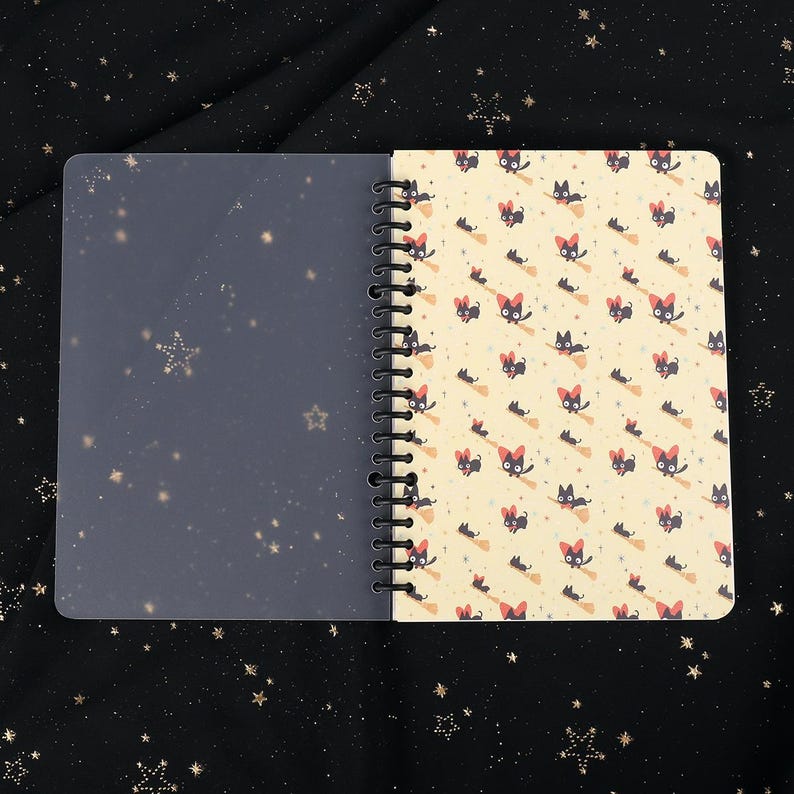 Puede incluir: Un cuaderno de espiral con una cubierta de pl&aacute;stico transparente y una cubierta amarilla con un patr&oacute;n repetido de gatos negros montando escobas. El cuaderno tiene una encuadernaci&oacute;n en espiral negra.