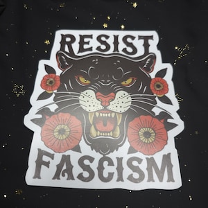 May include: A sticker featuring a black panther with yellow eyes and red flowers on either side. The words "RESIST FASCISM" are printed above and below the panther in bold, white letters. The background is black with gold flecks.