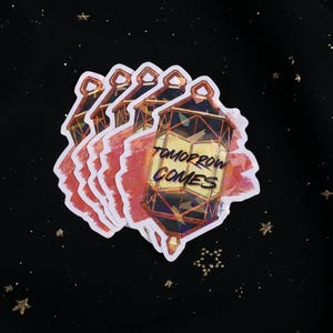 The Holographic lumina converter sticker with the text "Tomorrow Comes" on top. Behind the keychain is a red paint swipe.