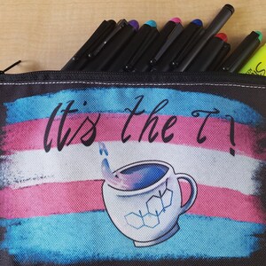 Puede incluir: Estuche negro con cremallera, con el texto "It's the T!" sobre un gr&aacute;fico de una taza de caf&eacute; con una estructura molecular. El estuche contiene bol&iacute;grafos negros y un resaltador amarillo.