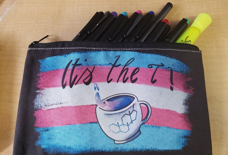 Puede incluir: Estuche negro con cremallera, con el texto "It's the T!" sobre un gr&aacute;fico de una taza de caf&eacute; con una estructura molecular. El estuche contiene bol&iacute;grafos negros y un resaltador amarillo.