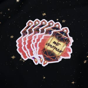 The Holographic lumina converter sticker with the text "We Continue" on top. Behind the keychain is a red paint swipe.