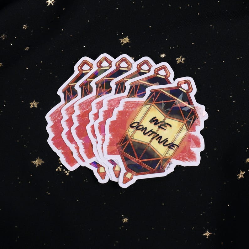The Holographic lumina converter sticker with the text "We Continue" on top. Behind the keychain is a red paint swipe.
