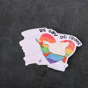 A sticker with a goose holding a knife on top of a rainbow heart, holding a knife in it's beak. The top says "Be Gay Do Crime"