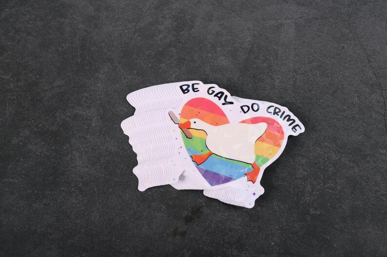 A sticker with a goose holding a knife on top of a rainbow heart, holding a knife in it's beak. The top says "Be Gay Do Crime"