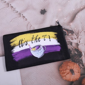 Puede incluir: Bolsa negra con cremallera con el texto "It's the T!" y un dise&ntilde;o de taza de t&eacute;. El dise&ntilde;o incorpora los colores de la bandera no binaria: amarillo, blanco y morado. La bolsa rectangular parece ser de tela.