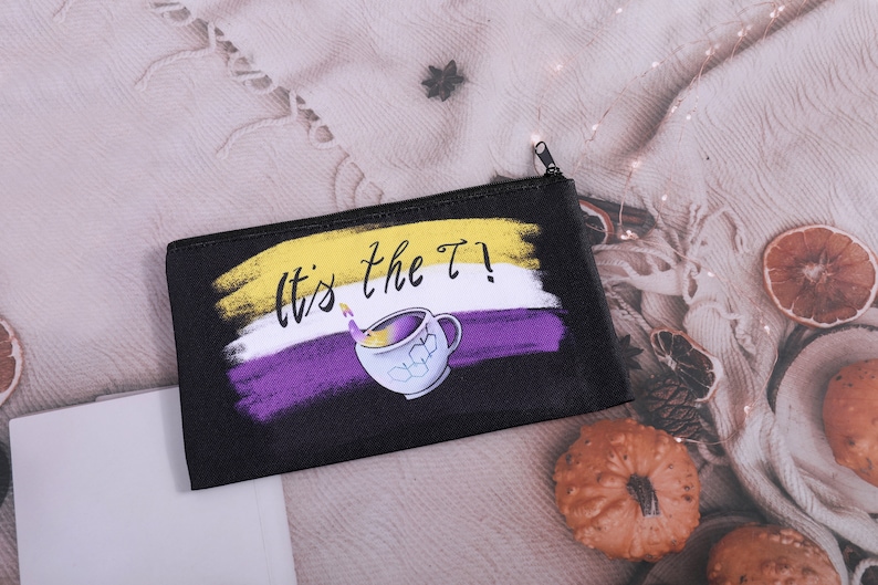 Puede incluir: Bolsa negra con cremallera con el texto "It's the T!" y un dise&ntilde;o de taza de t&eacute;. El dise&ntilde;o incorpora los colores de la bandera no binaria: amarillo, blanco y morado. La bolsa rectangular parece ser de tela.