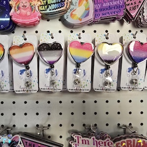 以下が含まれることがあります： プライドフラッグをテーマにしたハート型のバッジホルダー8個のディスプレイ。各バッジホルダーは透明なレジン製で、シルバーの引き込み式バッジリールが付いています。バッジホルダーは、各カードに「AnarchyArt Etsy.com」の文字が印刷された白いペグボードに表示されています。