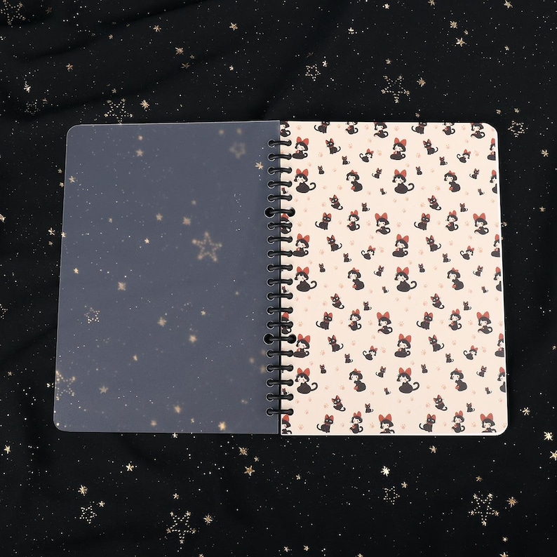 Puede incluir: Un cuaderno de espiral con una cubierta de pl&aacute;stico transparente y una cubierta de color crema con un patr&oacute;n repetido de gatos negros con lazos rojos.