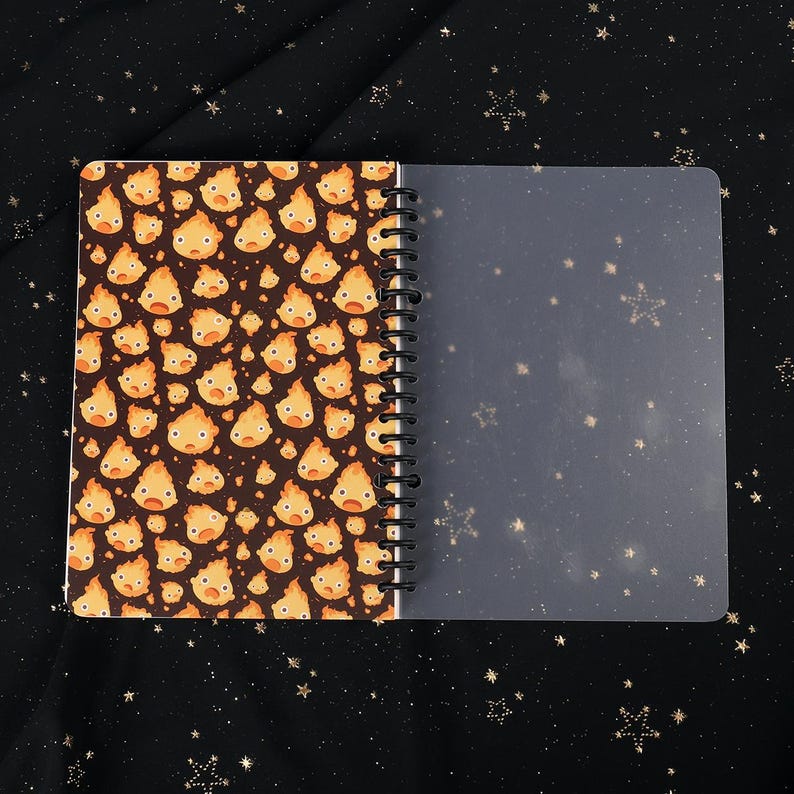 Puede incluir: Un cuaderno de espiral con una cubierta negra que presenta un patr&oacute;n repetido de sprites de fuego amarillos y naranjas de dibujos animados. El cuaderno tiene una cubierta de pl&aacute;stico transparente.