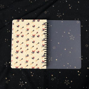 Puede incluir: Un cuaderno de espiral con una cubierta amarilla con un patr&oacute;n repetido de gatos negros montando escobas. El cuaderno tiene una cubierta de pl&aacute;stico transparente.